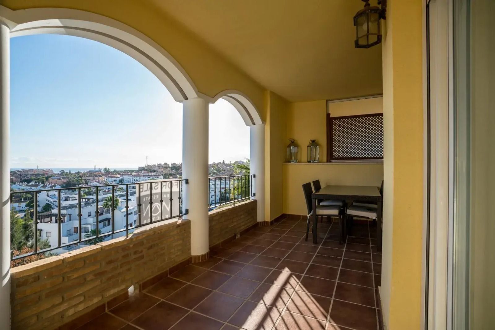 Covered balcony with arches, tiled floor, and a table overlooking a waterfront city view