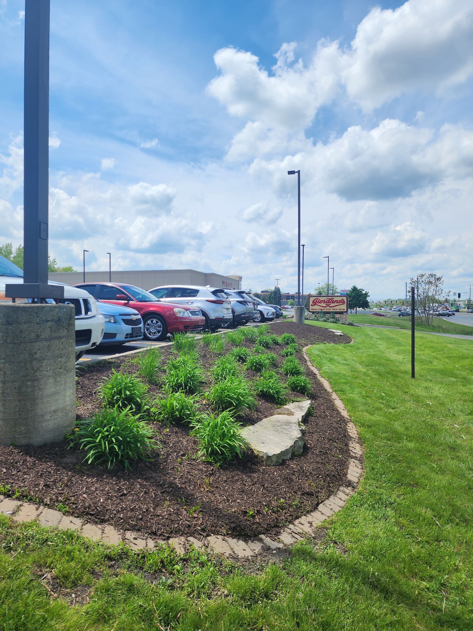 Mulched flower bed with rocks beside a parking lot under a cloudy blue sky