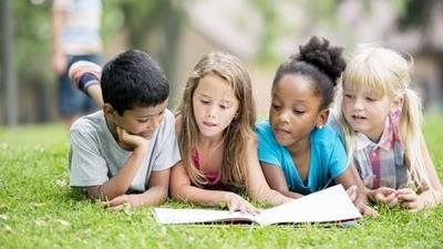 Four children lying on grass, reading an open book together outdoors.