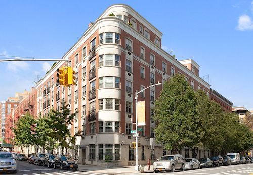 Red-brick corner apartment building on a city street with trees and traffic lights under a blue sky