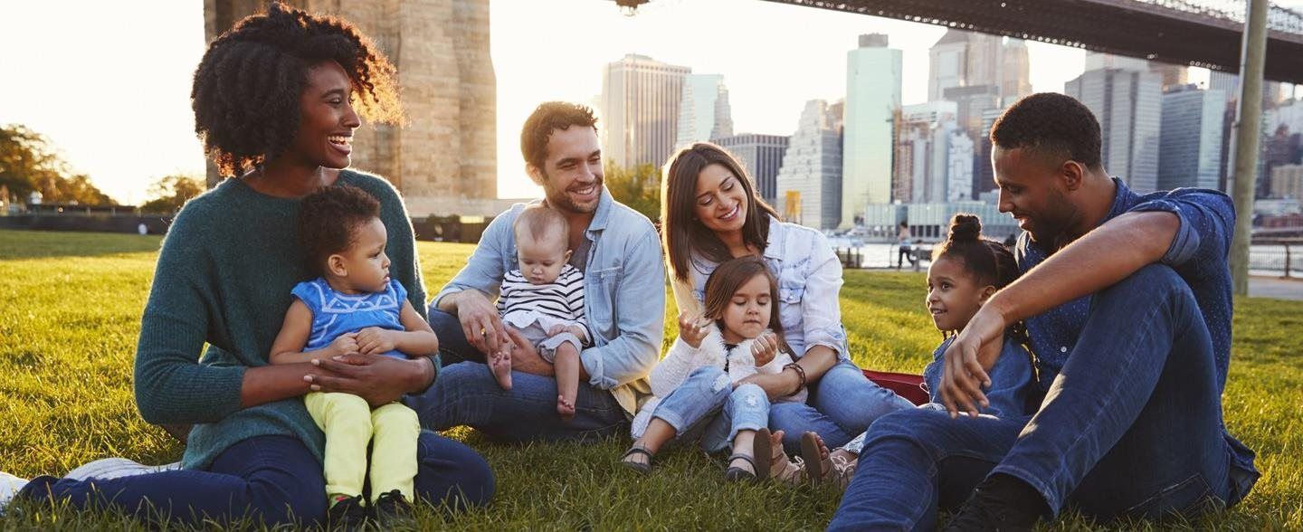 Friends sitting on grass with babies, talking and smiling in a city park at sunset