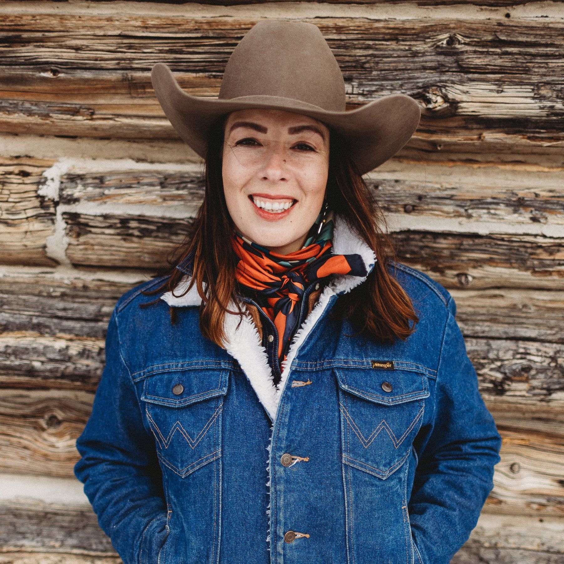 Woman in cowboy hat and denim jacket smiles against a wood-paneled wall.