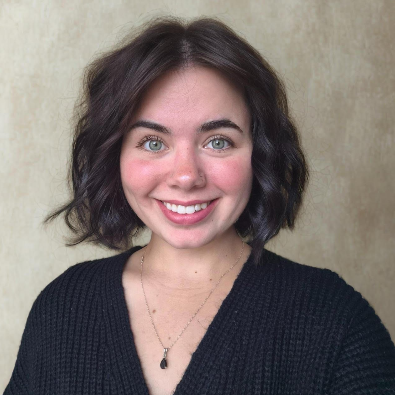 Woman with short dark hair, smiling, wearing a black sweater and a necklace. Neutral background.