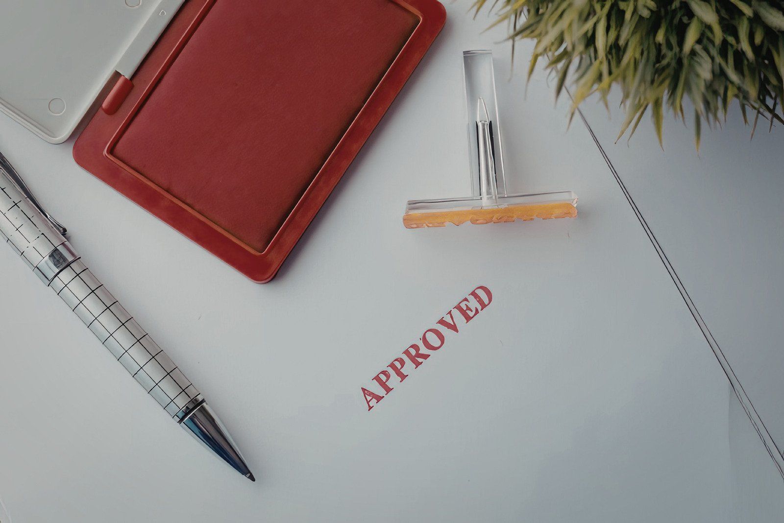 Red notebook, pen, and stamp with “APPROVED” on a white desk.