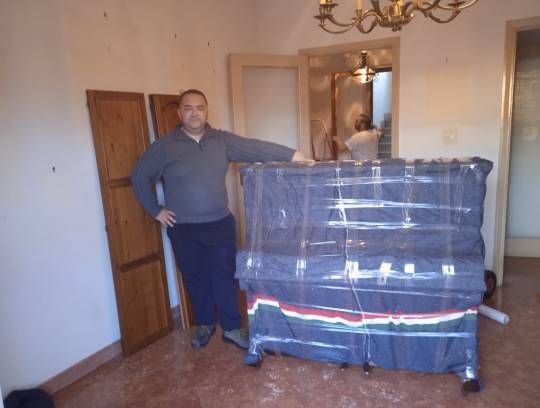 Un uomo in piedi accanto a un pianoforte imballato in una stanza. Porte marroni e un ingresso con un'altra persona visibile.