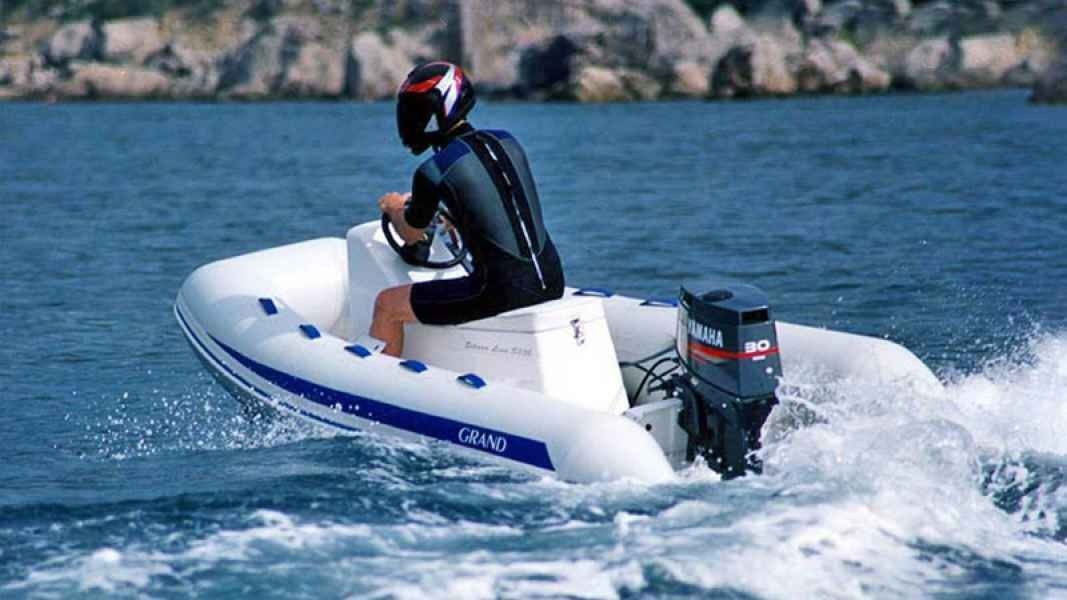Person riding a small white speedboat on blue water, leaving a wake near a rocky shoreline.