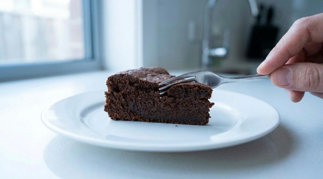 A close up of a fork cutting into a dense slice of chocolate mud cake with a kitchen faucet in the background.
