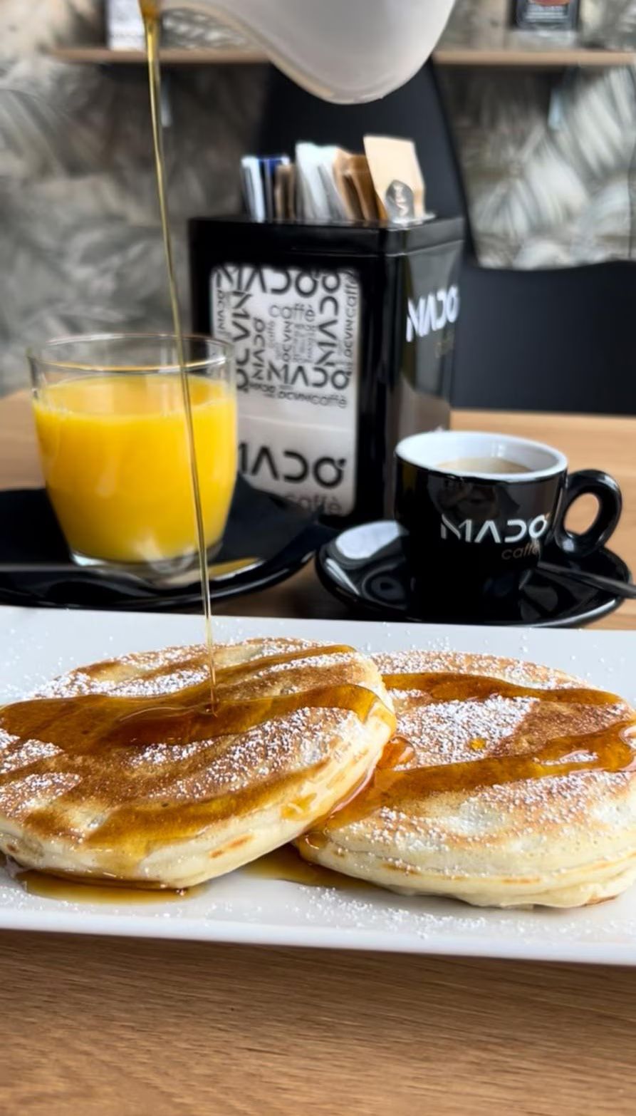Sciroppo versato sui pancake su un piatto, con caffè e succo di frutta su un tavolo.