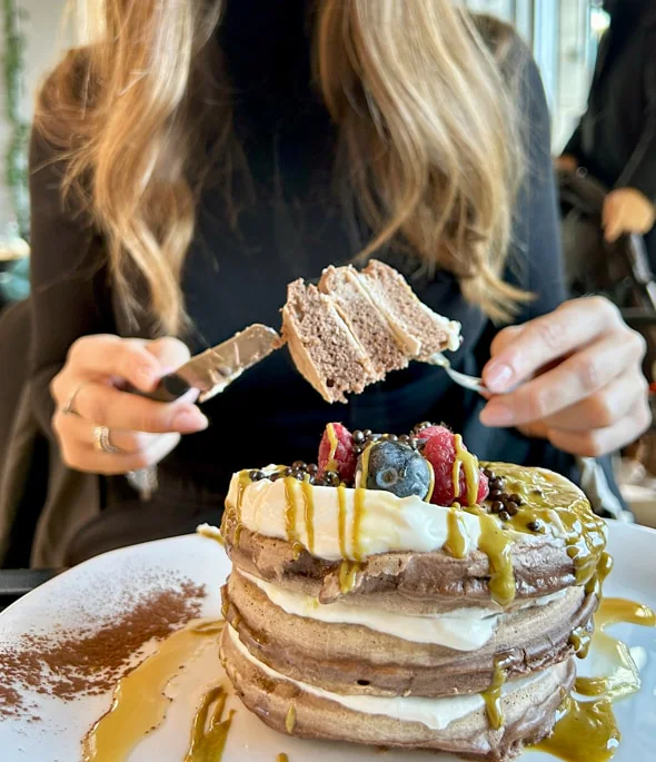 Donna che mangia una pila di pancake con frutta, panna e salsa. Ambiente interno.