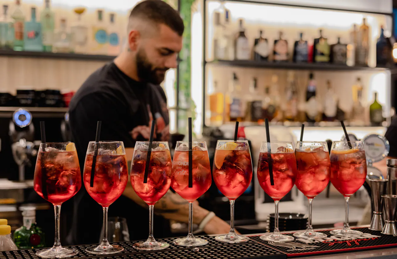Il barista prepara una fila di cocktail rossi in un bar, concentrandosi sui drink e sullo sfondo.