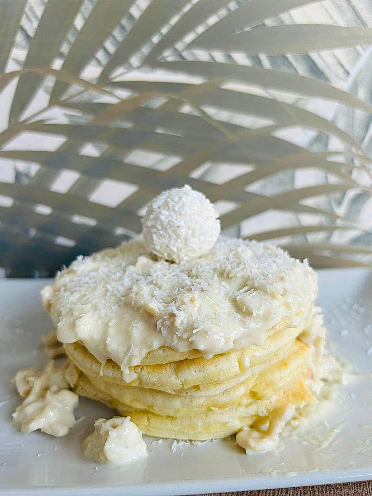 Pila di pancake con glassa bianca, scaglie di cocco e una pallina di cocco su un piatto bianco.