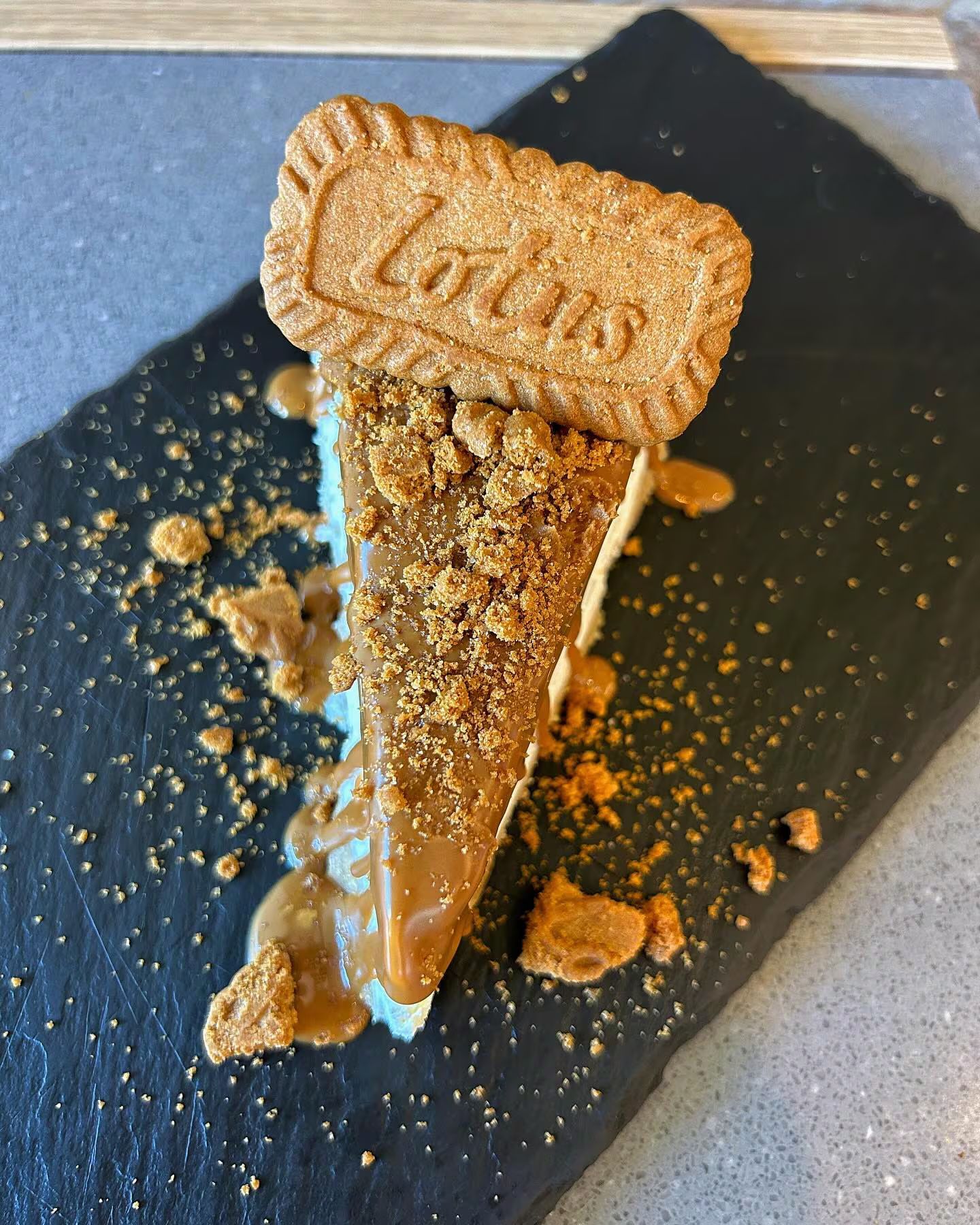 Fetta di cheesecake, guarnita con briciole di biscotti e salsa al caramello, e un biscotto Lotus, su una lavagna nera.