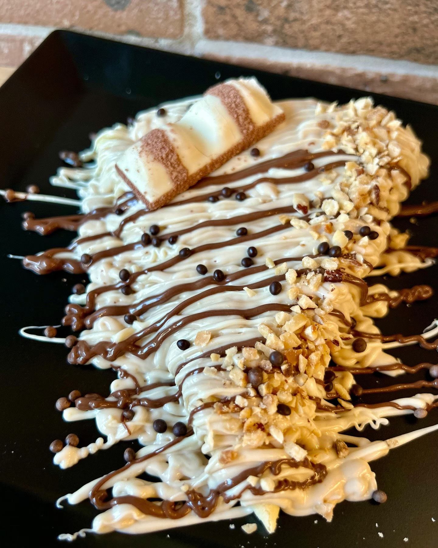 Crêpe condita con cioccolato e besciamella, guarnita con Kinder Bueno, noci e granella di cioccolato, servita su un piatto nero.