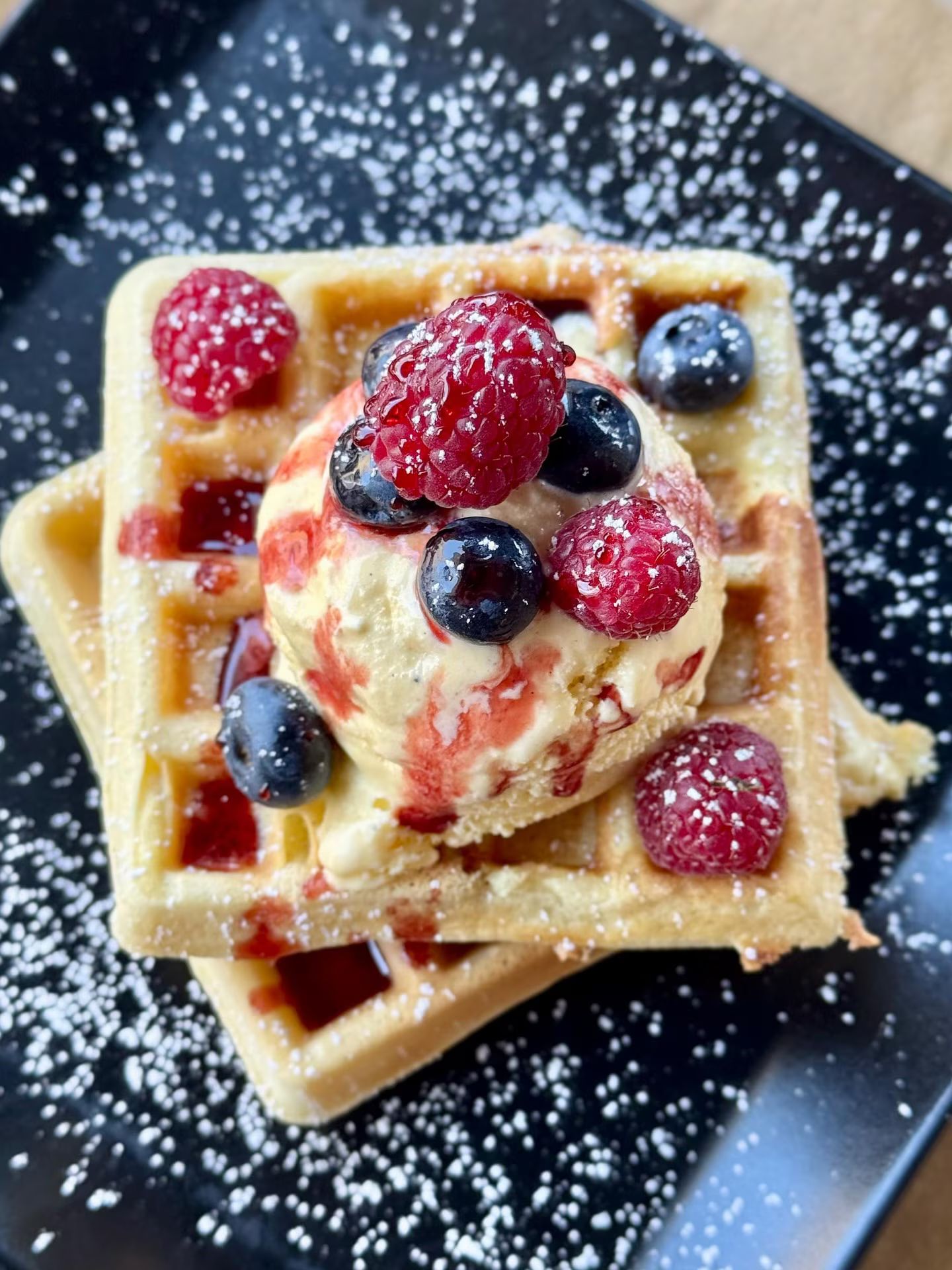 Waffle ricoperti di gelato, lamponi e mirtilli, spolverati con zucchero a velo, su un piatto nero.