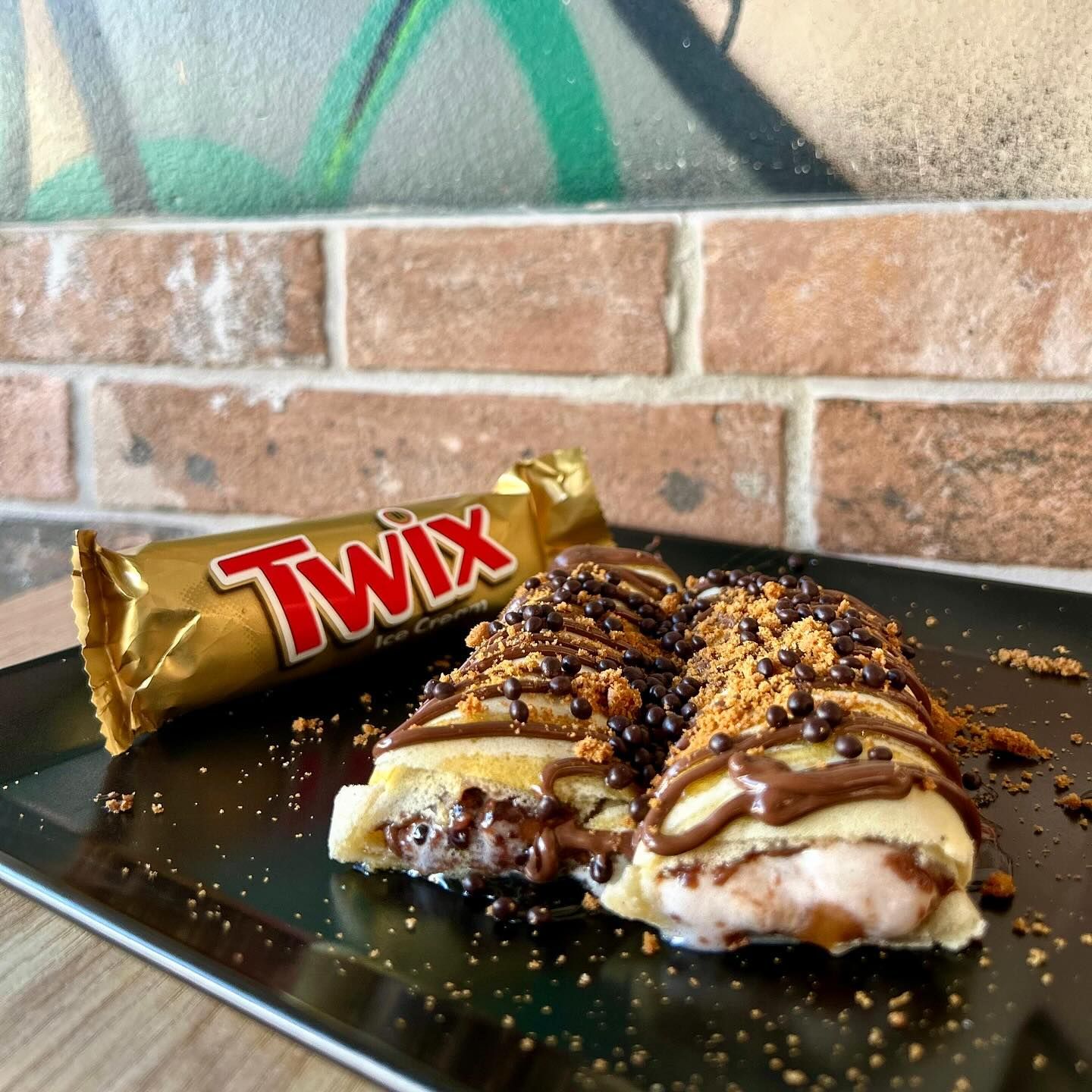 Crêpes ripiene di Twix ricoperte di cioccolato e guarnite con biscotti sbriciolati, accanto a una barretta di Twix su un piatto nero.