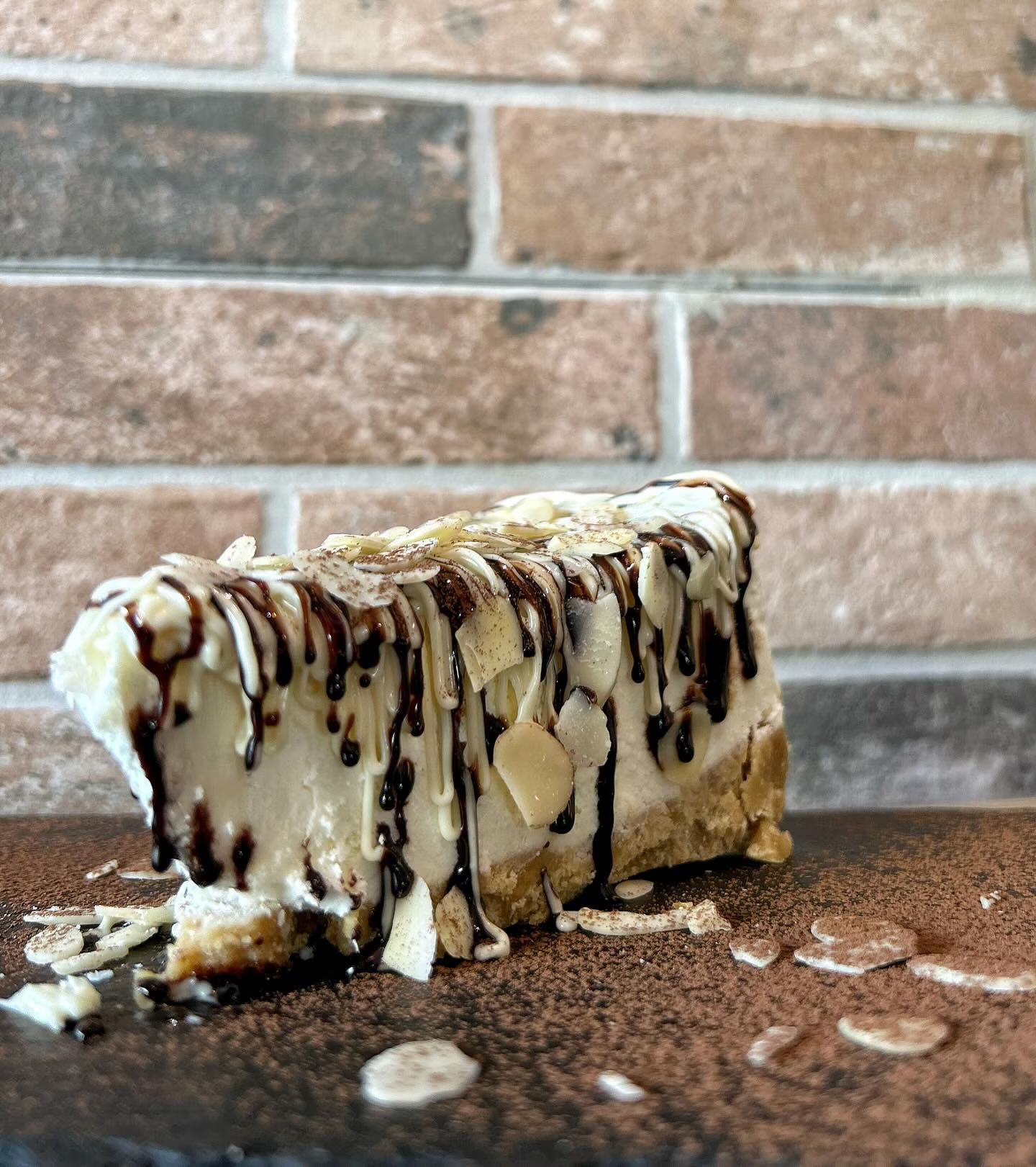 Fetta di cheesecake con glassa di cioccolato e mandorle su una superficie marrone, sfondo mattone.