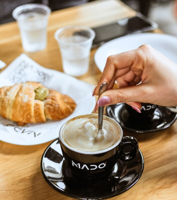 Una mano che mescola un cappuccino schiumoso in una tazza nera Mado, un croissant e dei bicchieri d'acqua su un tavolo.