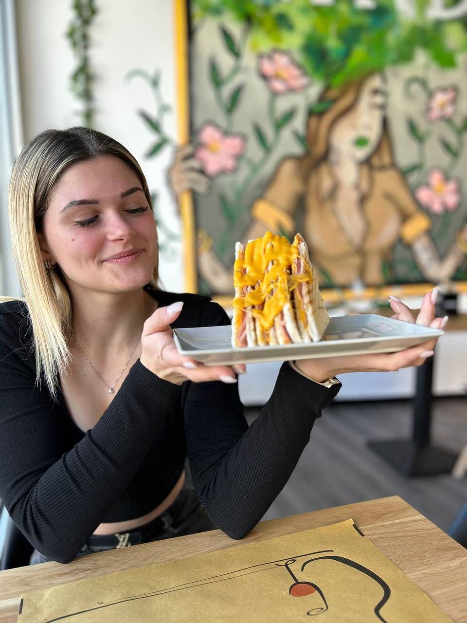 Donna sorridente che tiene in mano un grande panino al formaggio in un bar, con un dipinto sullo sfondo.