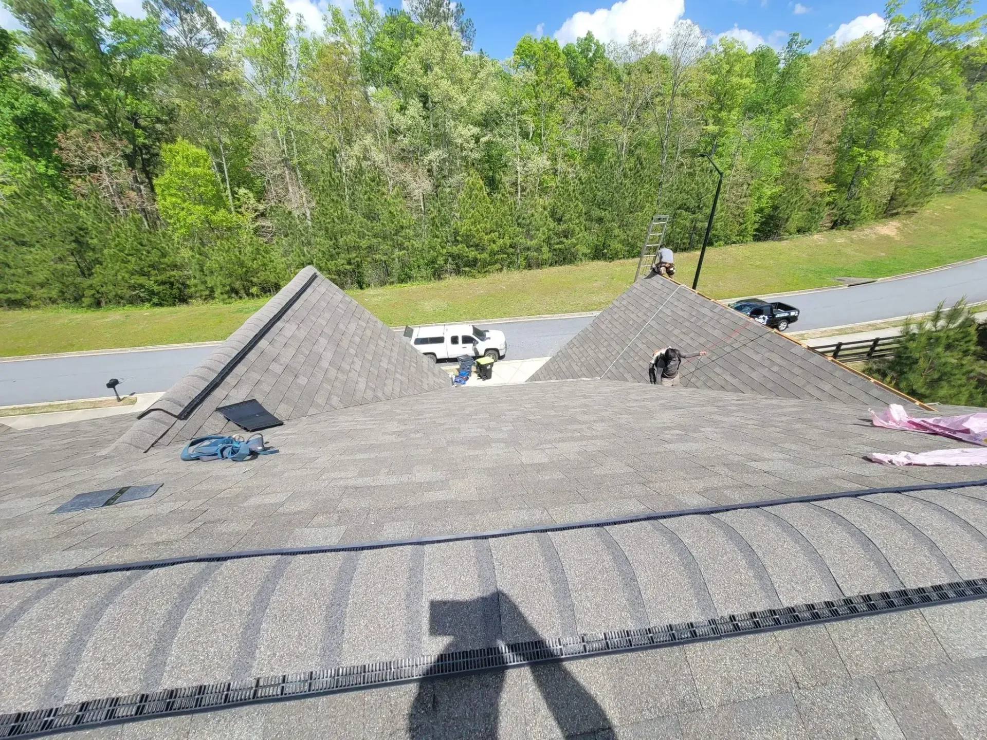 Workers on a rooftop with piles of gray roofing material, trees and road in the background.