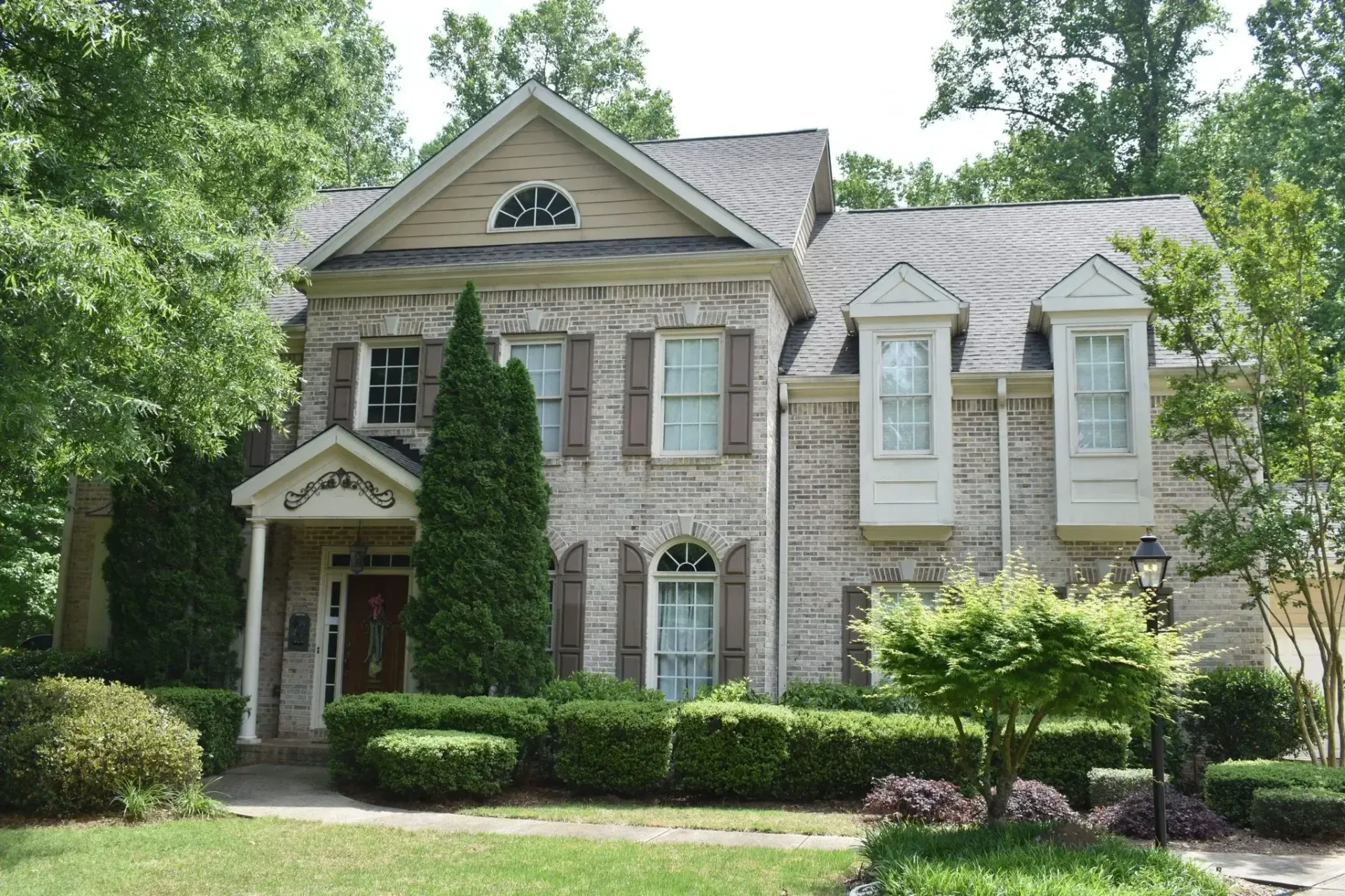 Two-story brick house with shutters, surrounded by greenery.