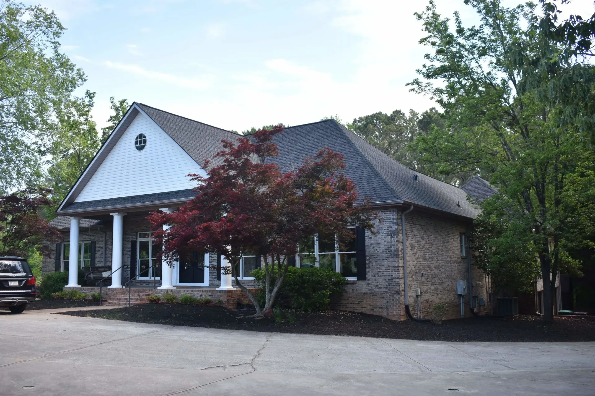 A one-story brick building with a white porch, trees, and a parked car.