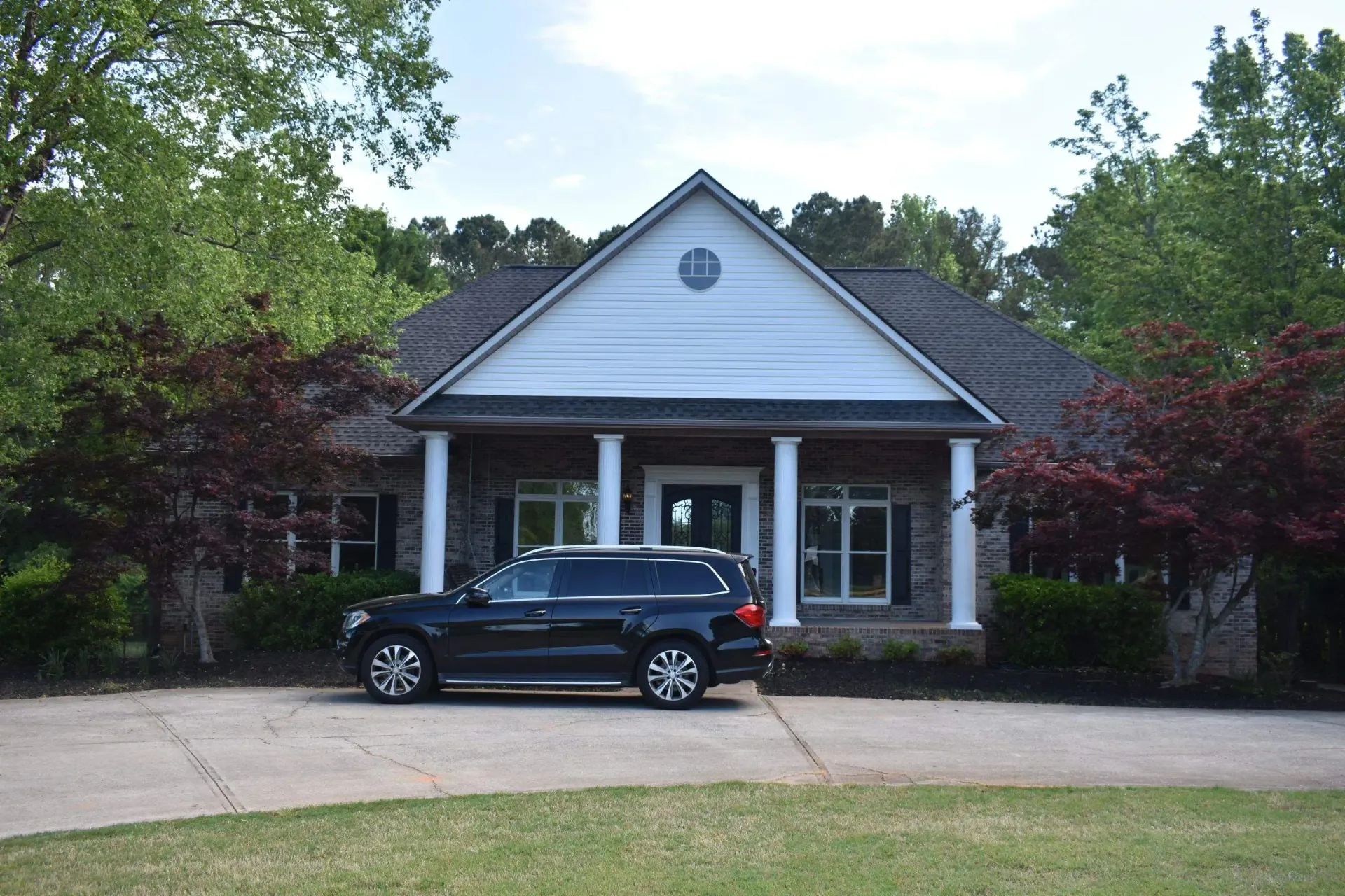 Black SUV parked in front of a brick house with white columns and a slanted roof. Green trees surround.