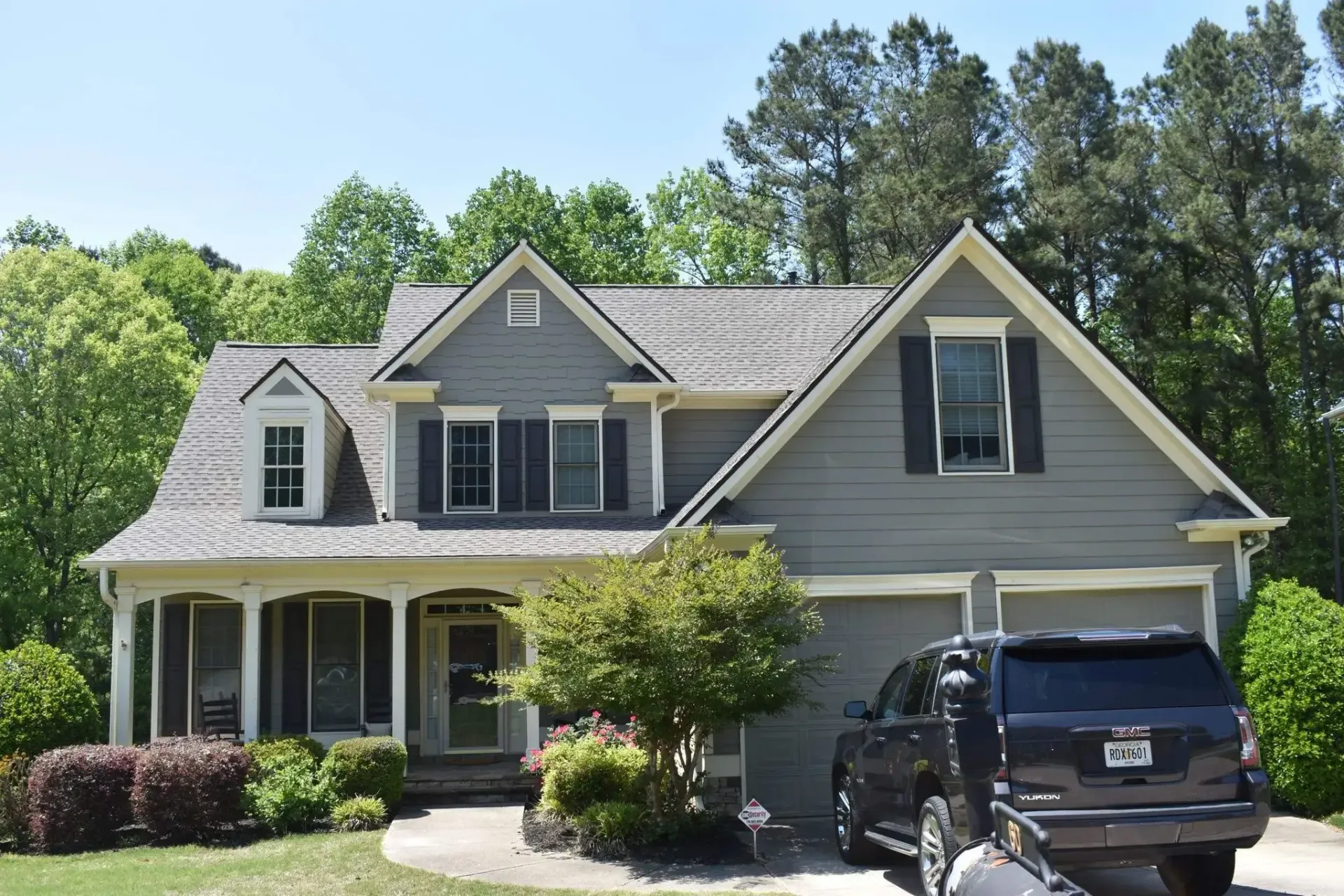 Gray two-story house with a car parked in the driveway, surrounded by green trees and bushes on a sunny day.