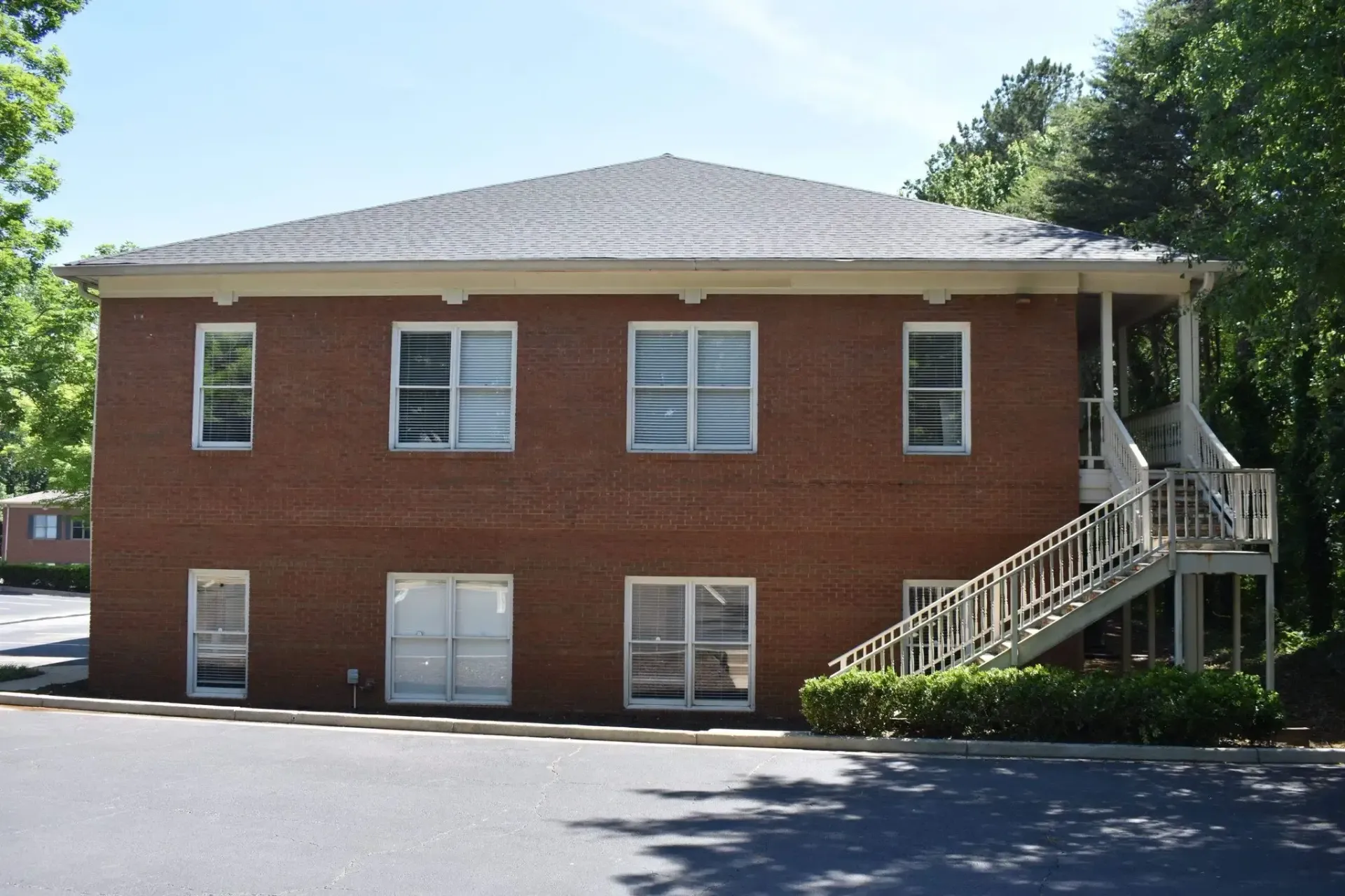 Two-story brick building with a white staircase on the side, surrounded by trees and a paved parking lot.