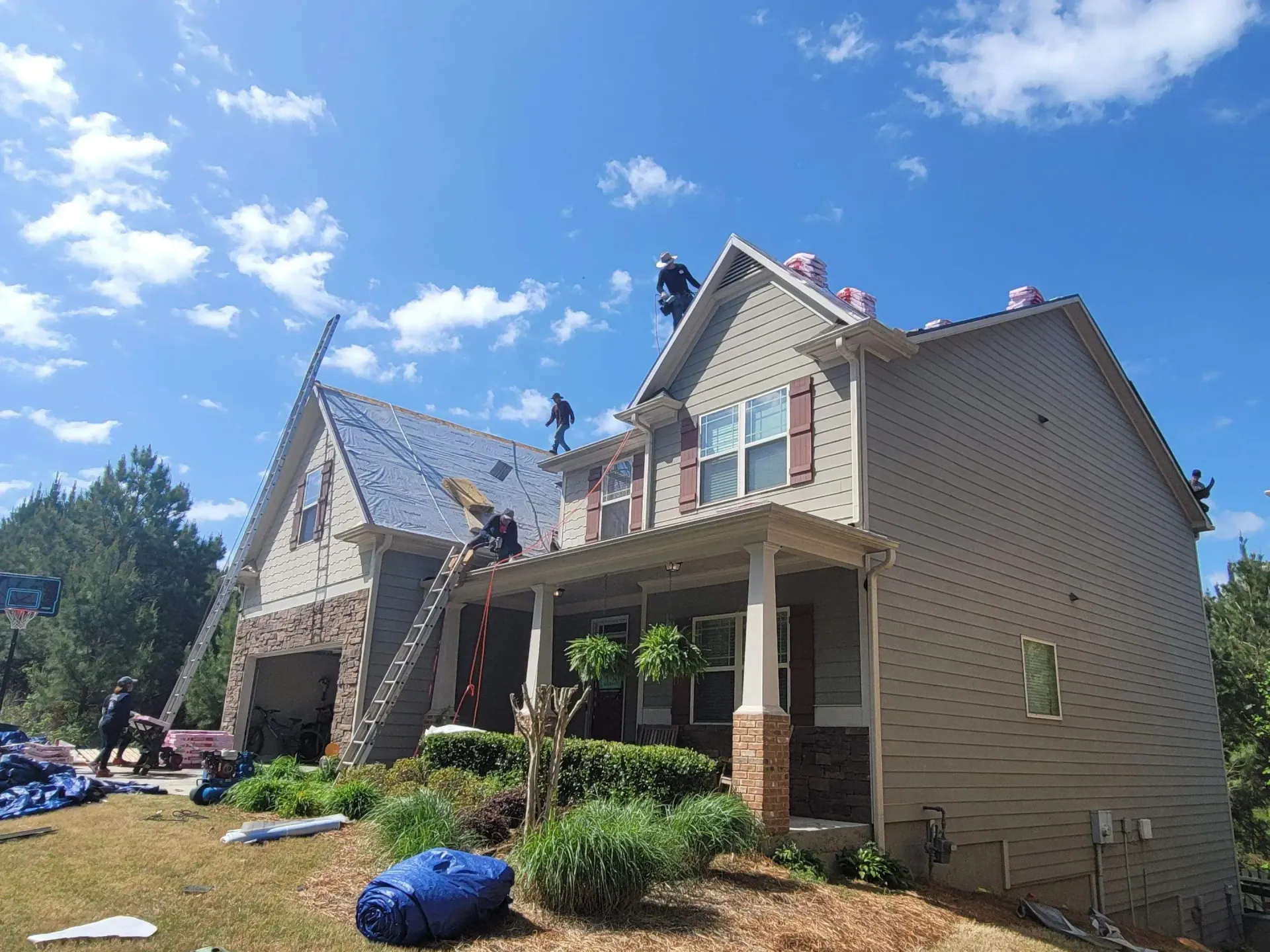 Roofers working on a two-story house on a sunny day. Blue tarps and a ladder are visible.