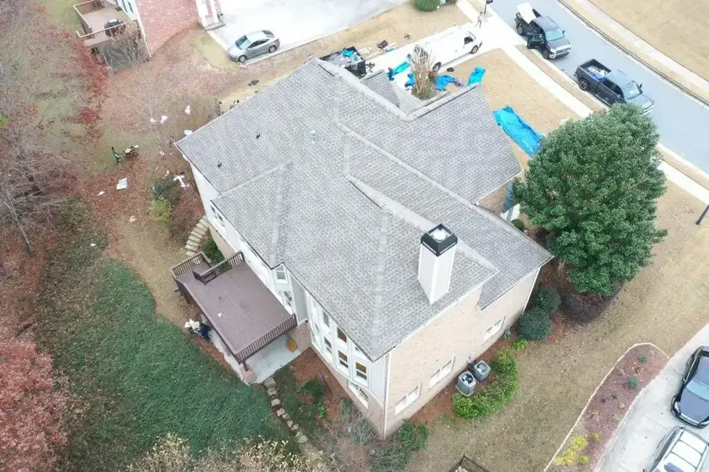 Aerial view of a house with a gray roof, deck, and surrounding yard. Cars are parked in the driveway.