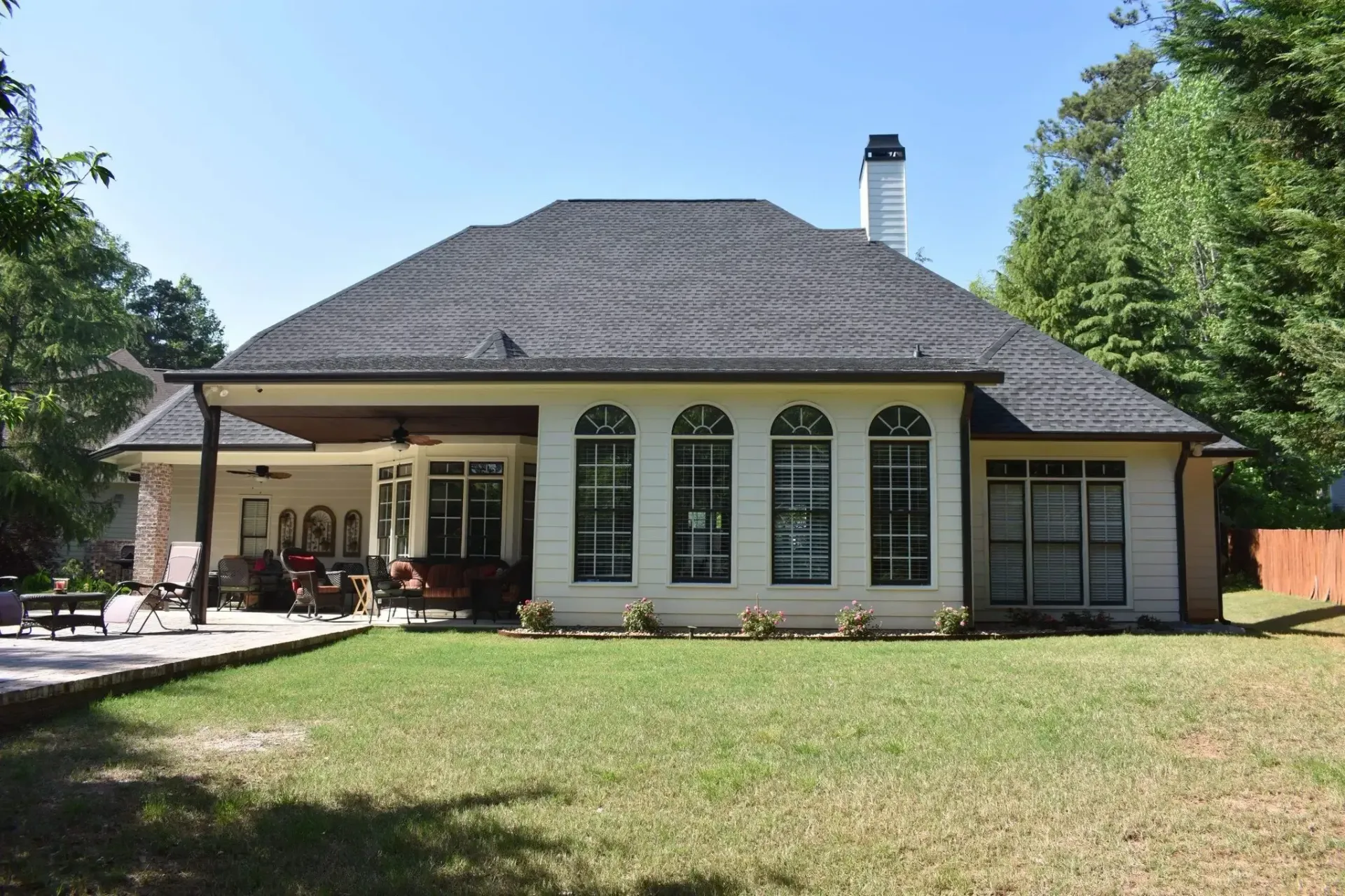 Back view of a light-colored house with a patio, arched windows, and a dark tiled roof on a sunny day.