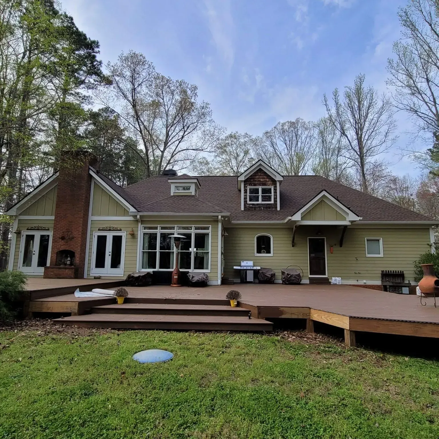 Tan house with brown deck and roof, surrounded by trees, on a sunny day.