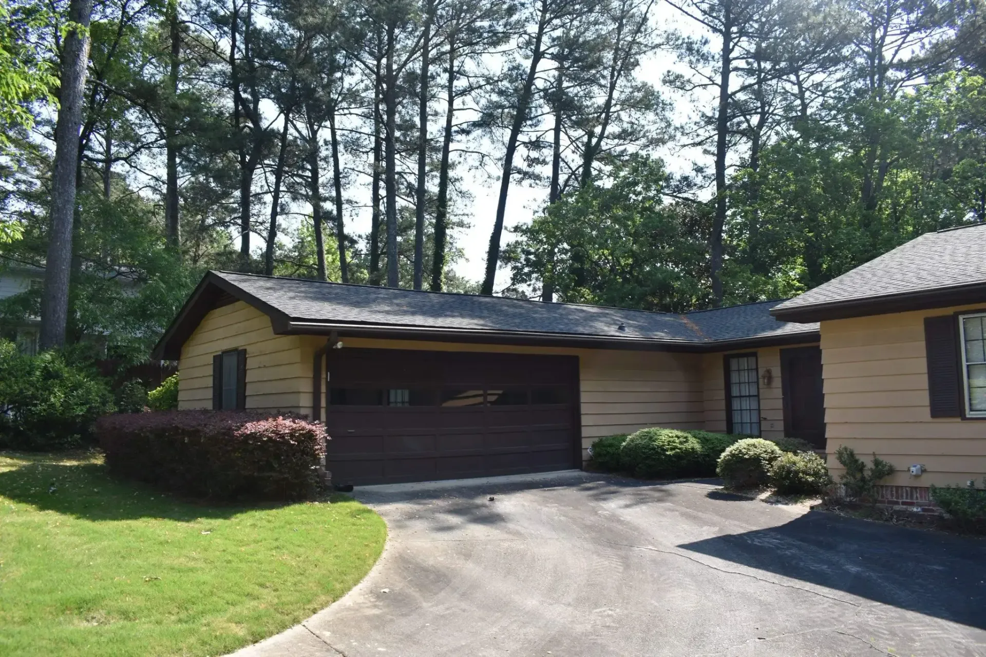House with brown garage door, tan siding, and green lawn, trees in the background.