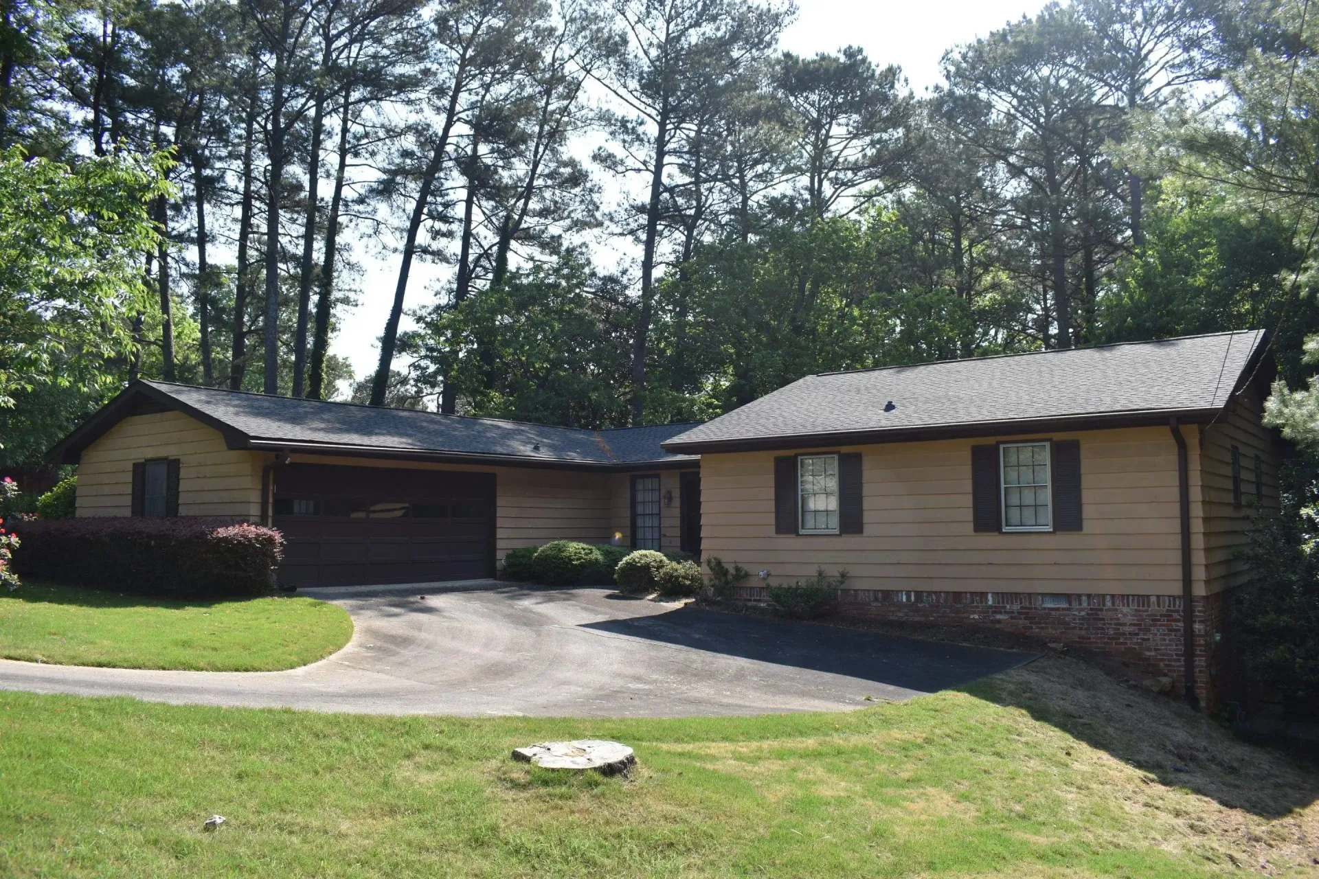 Tan house with a driveway, garage, and a separate structure to the right, set against a backdrop of trees.