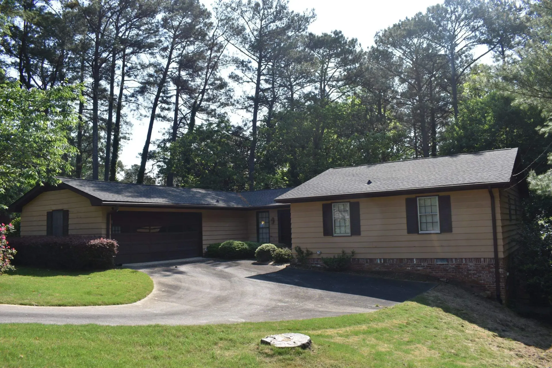 Tan ranch house with a dark garage door, driveway, and surrounding trees.