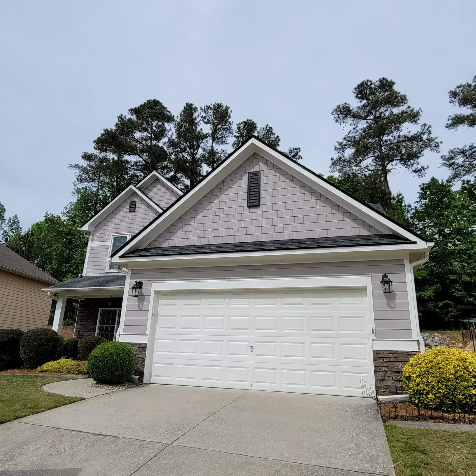 Two-story house with white garage door, gray siding and roof, and green bushes.