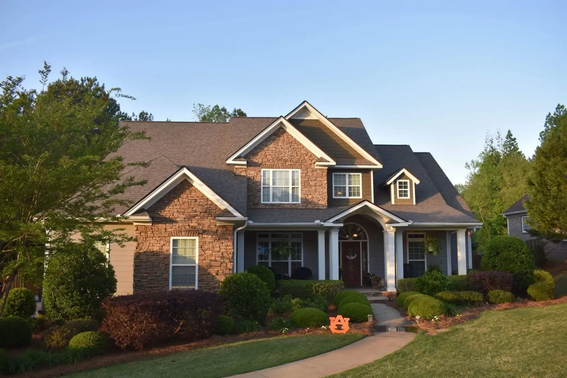 Two-story brick house with a porch and manicured landscaping, under a clear blue sky.