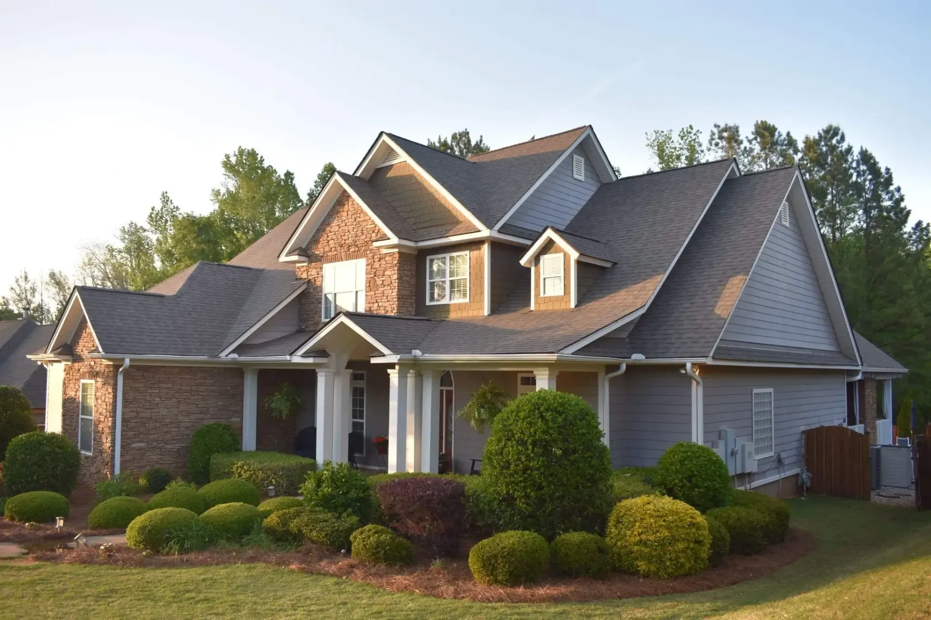 House with brown brick and siding, surrounded by bushes and greenery.