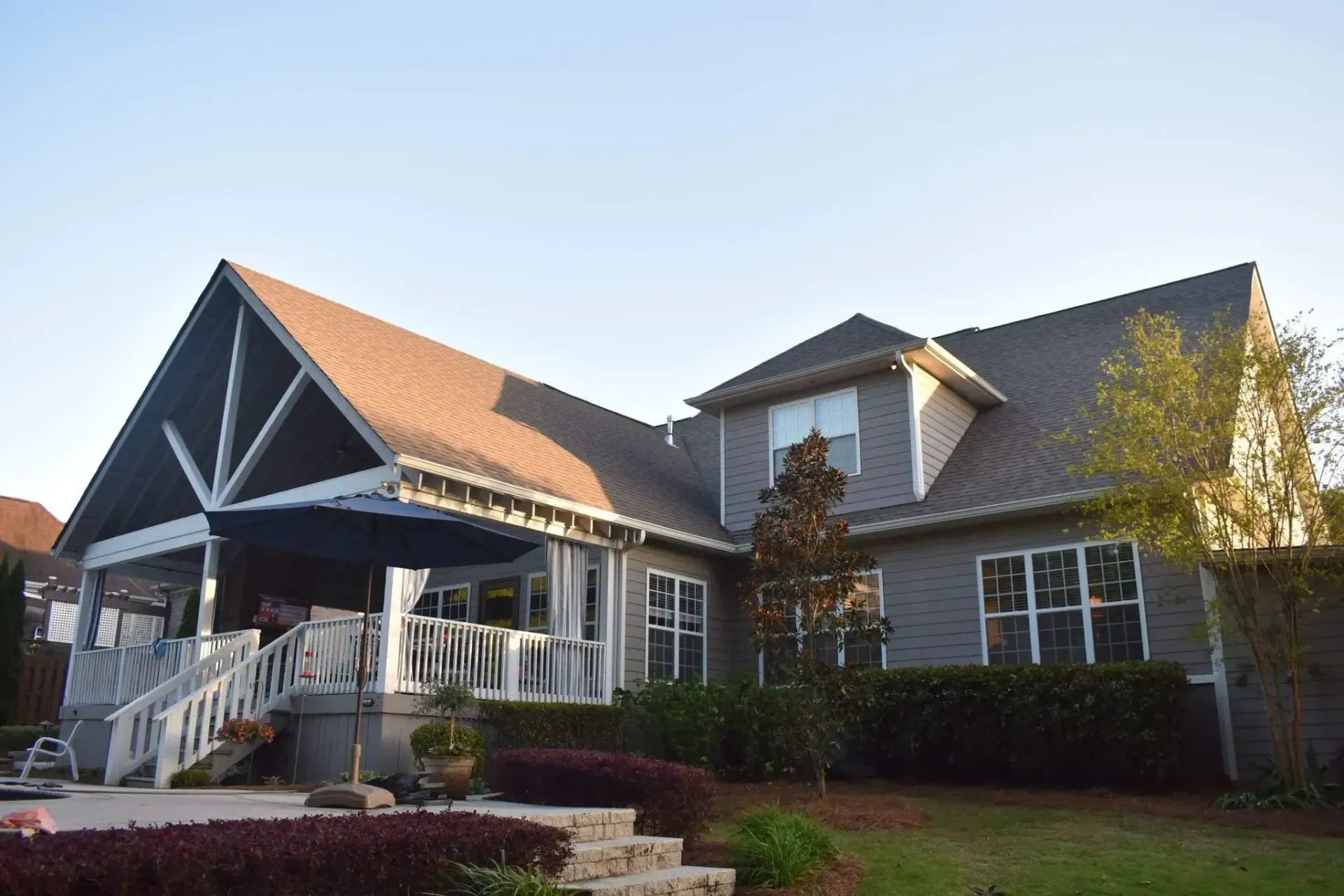 Gray house with a brown roof, a large porch, and lush landscaping against a blue sky.