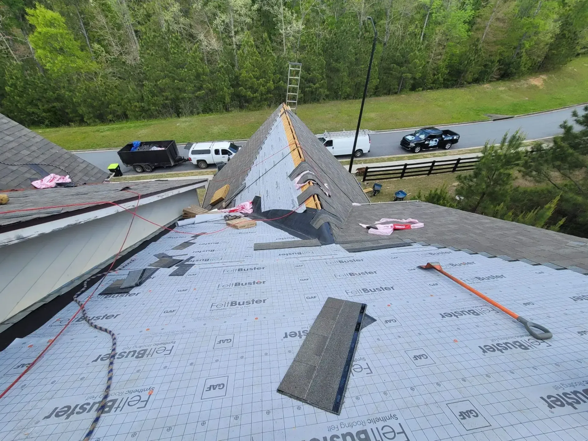 Roof partially covered with gray shingles, workers, vehicles, and forest background.