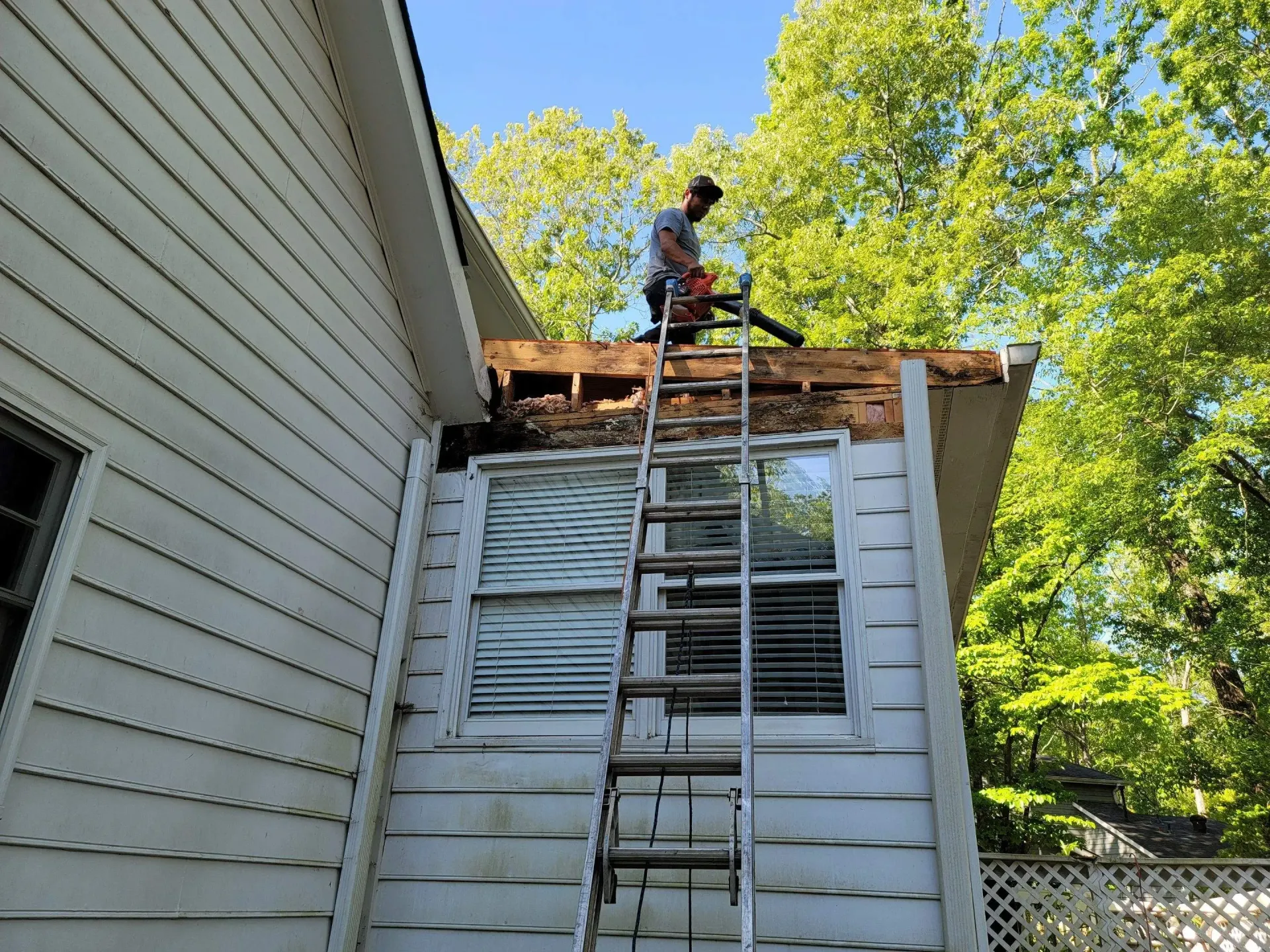 A man on a ladder blowing debris from a partially demolished roof. White house, green trees.