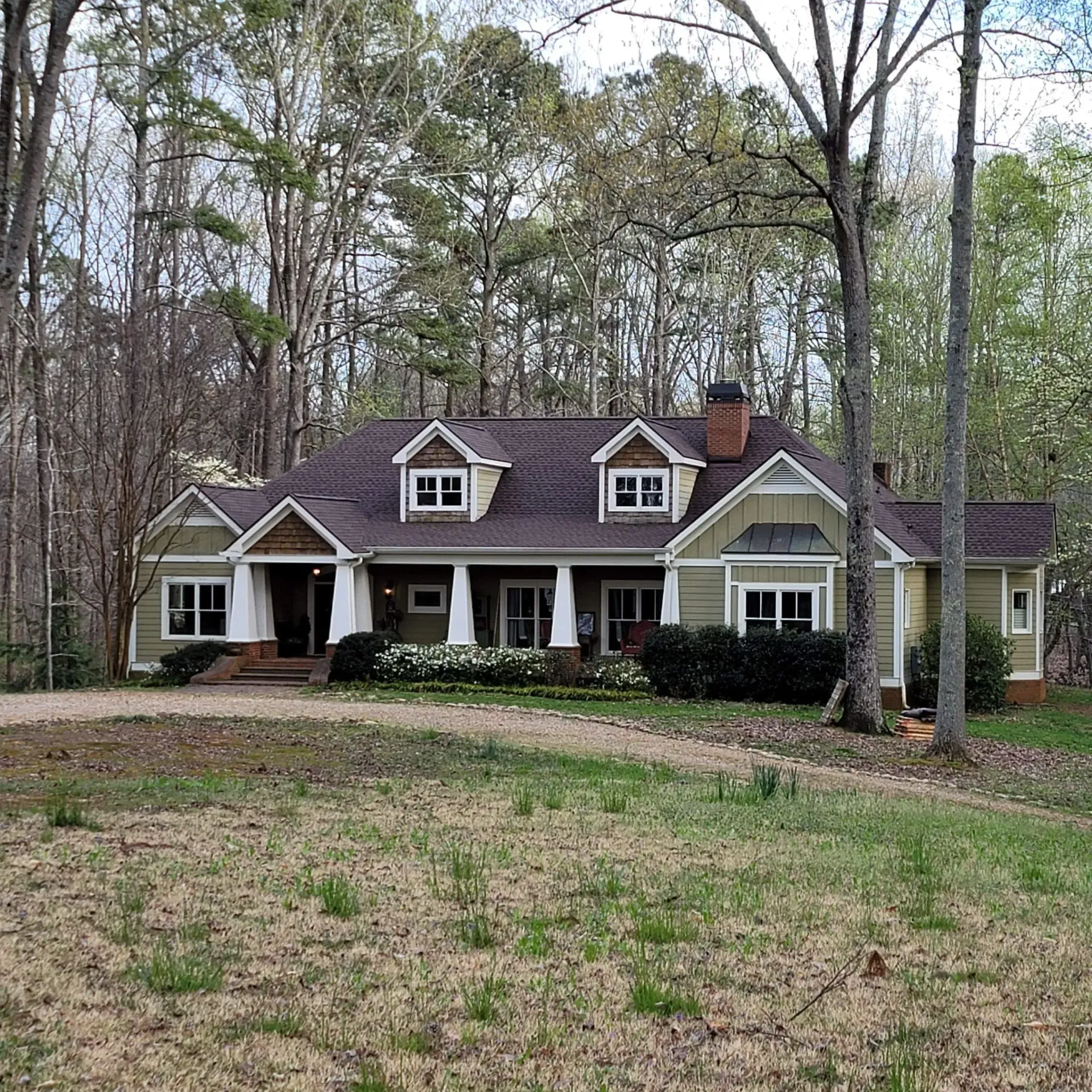 Green house with white trim, front porch, and brown roof in a wooded area.
