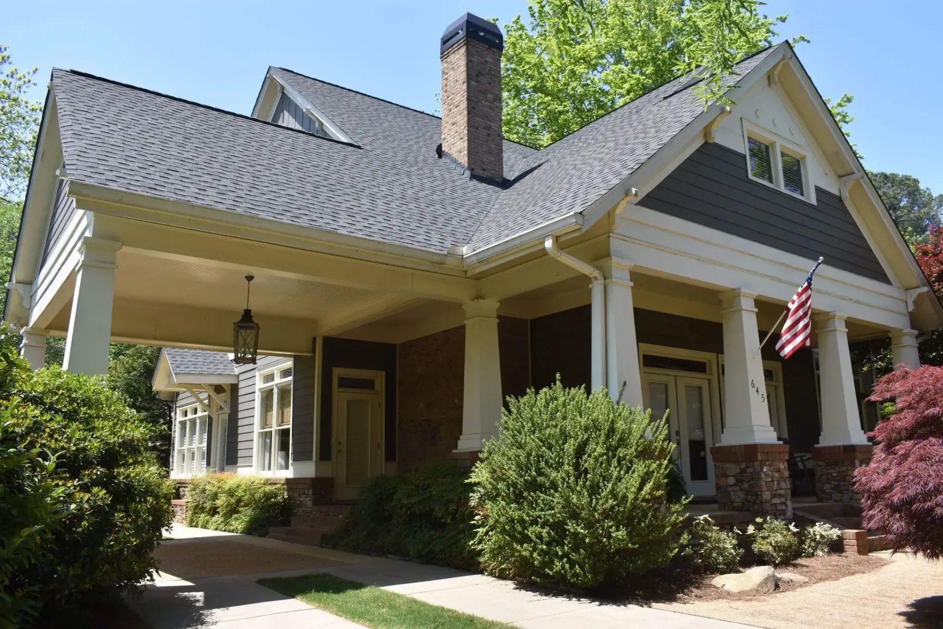 Cottage-style house with a porch, brick chimney, and US flag. Lush green and red bushes in the foreground.