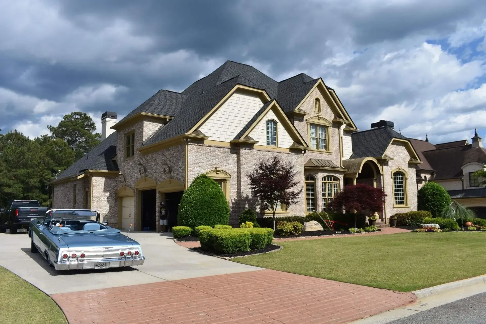 Large brick house with a blue convertible in the driveway. Overcast sky, manicured lawn.