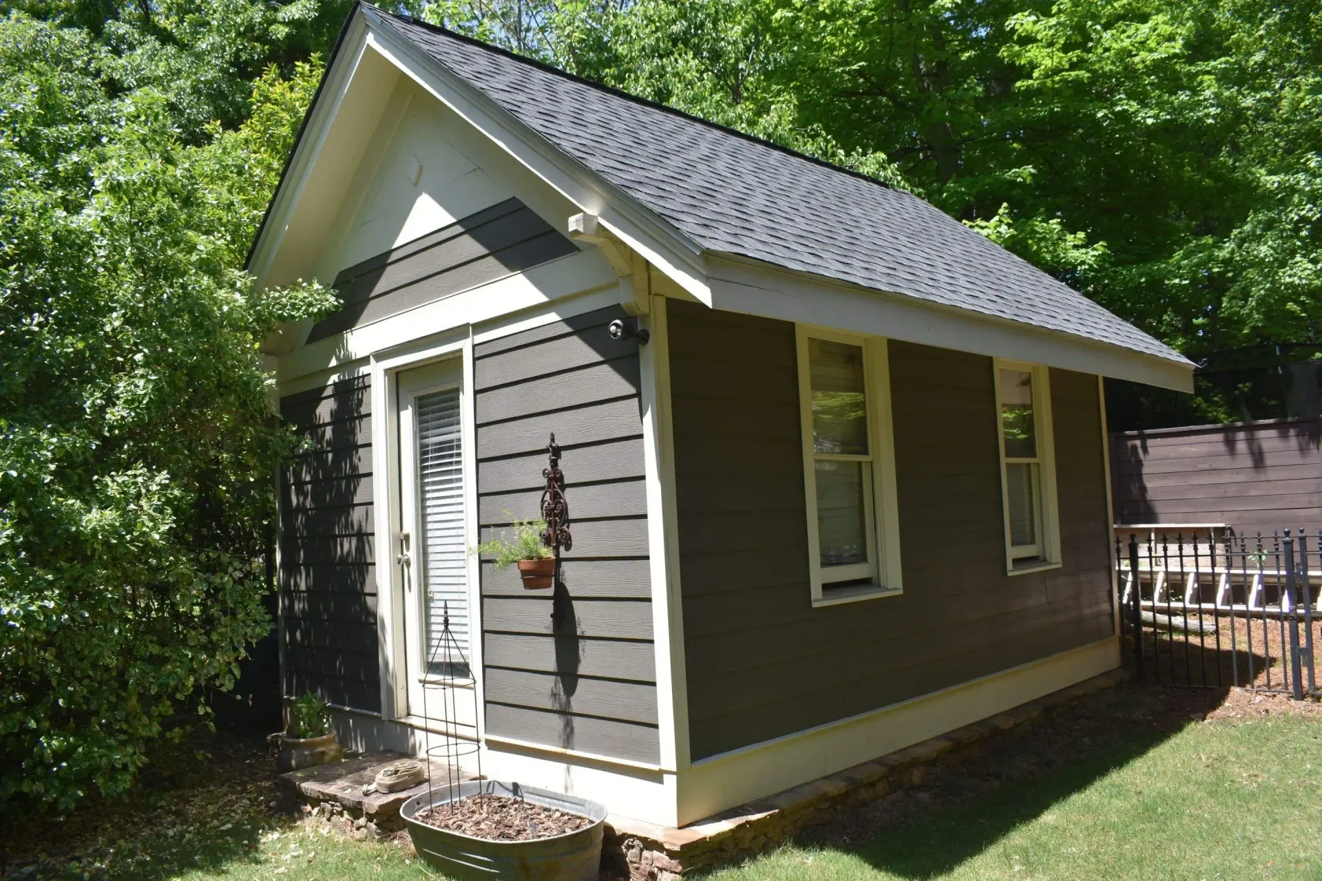 Small, dark-green building with white trim. Front door and two windows. Building is surrounded by trees and grass.