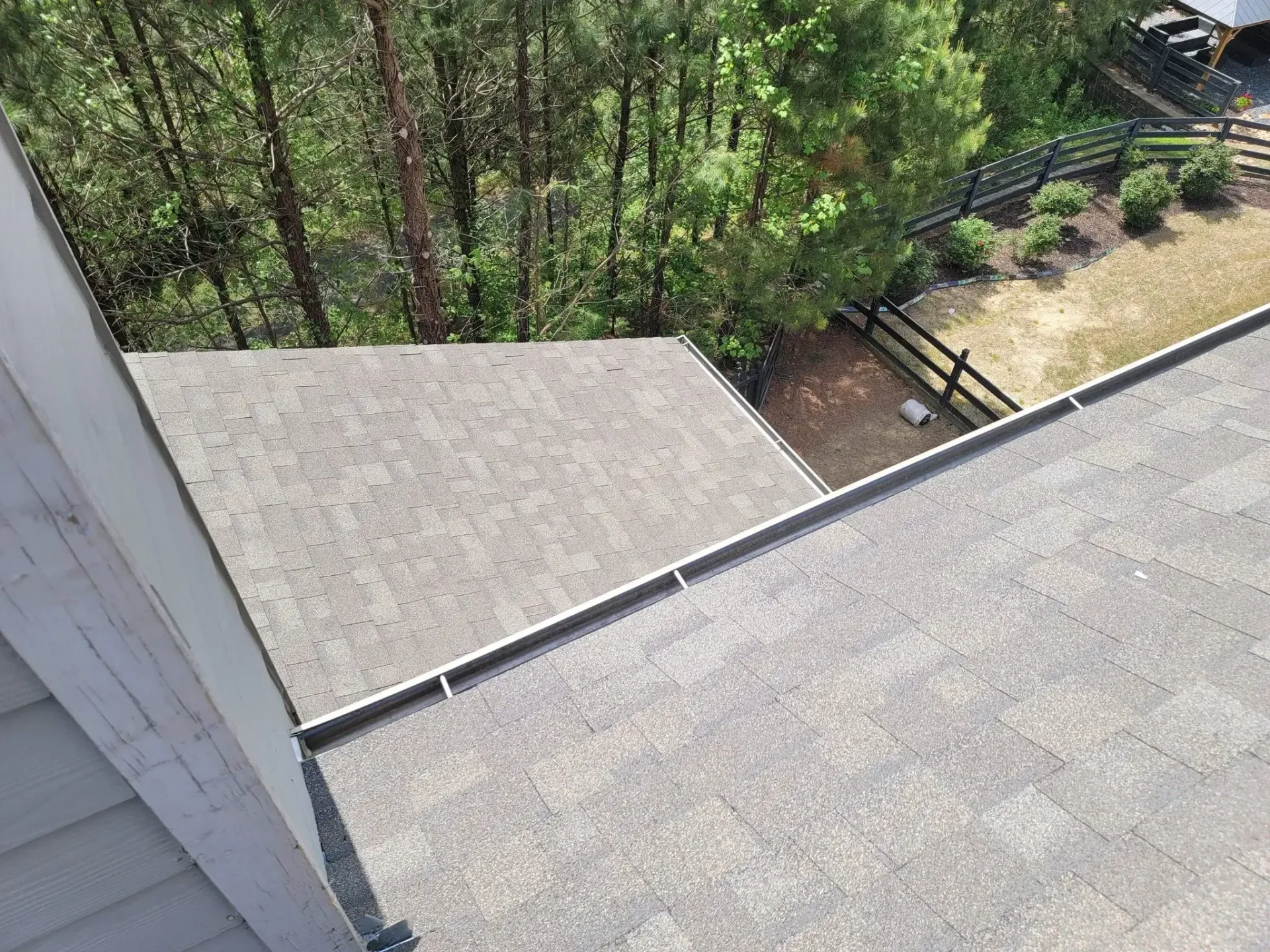 View of a gray asphalt shingle roof corner, next to siding and overlooking a wooded area and fence.