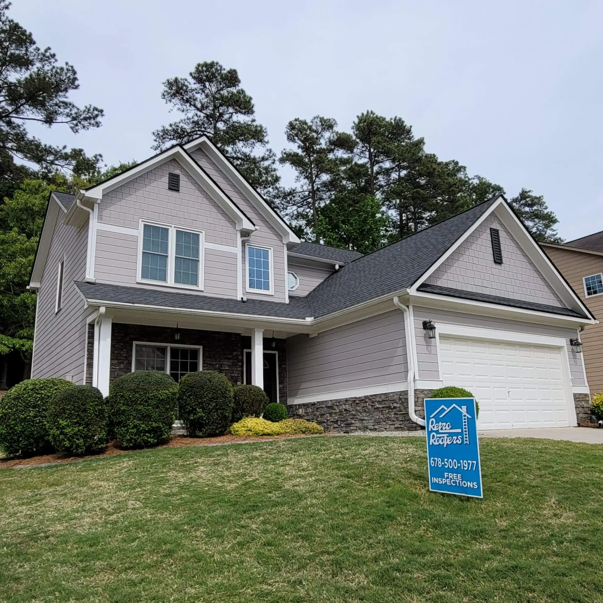 Two-story house with gray siding, dark roof, stone accents, and sign on a green lawn.