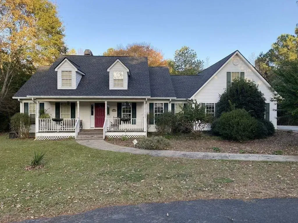 White house with dark roof, porch, red door, green shutters, and grassy yard.
