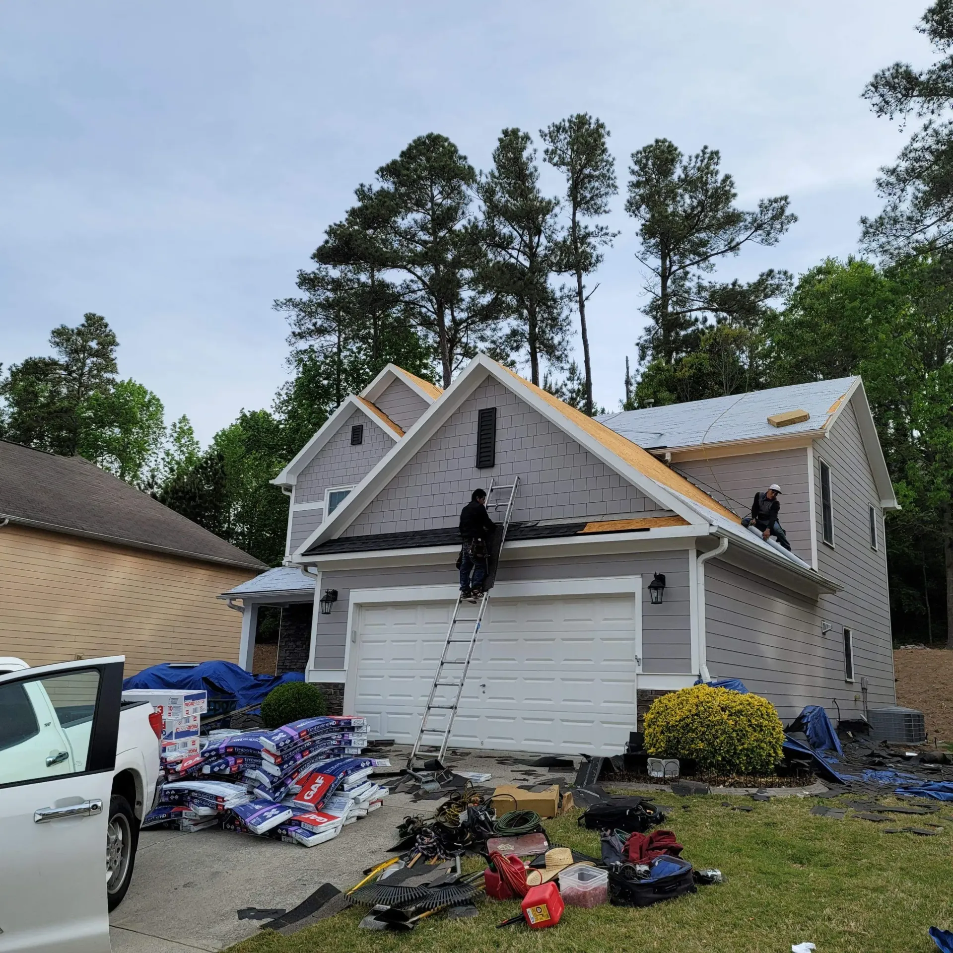 Roofers working on a two-story house, ladder propped against the building. Tools and materials are scattered on the ground.