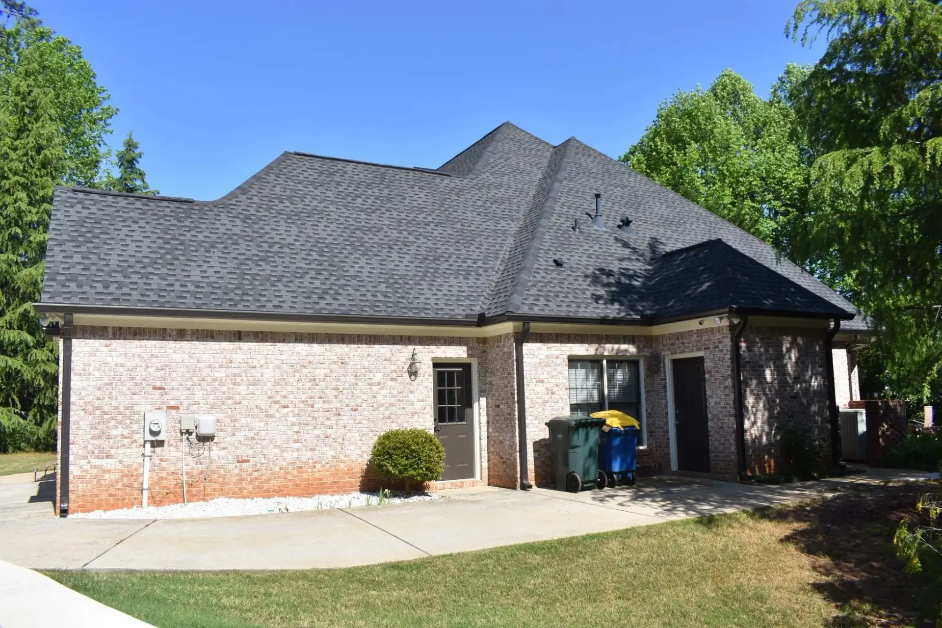 Tan brick house with a dark roof and green bushes on a sunny day.